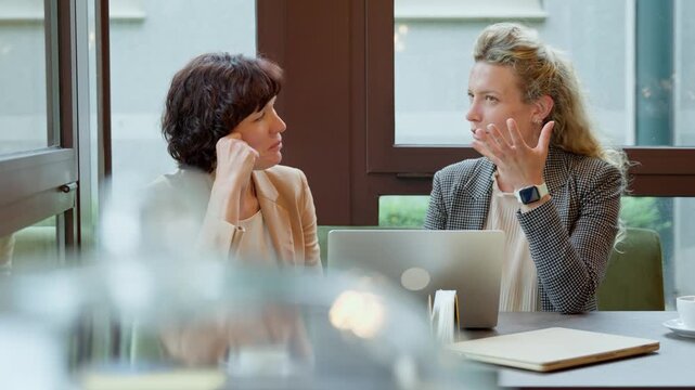 Female colleagues engaging in a friendly conversation at a cafe table. One businesswoman gestures while speaking as her partner listens intently