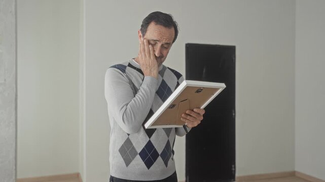 Man holding picture frame, hand on cheek in house, wearing argyle sweater, looking down at portrait; loss reflection grief.