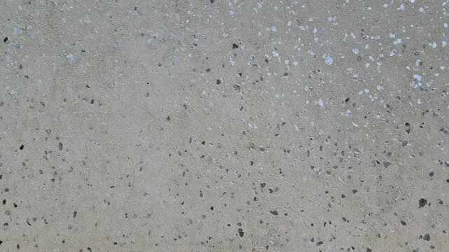 A flat, beige concrete or stone surface with a speckled texture, featuring scattered small grey, brown, and white mineral fragments throughout the material.