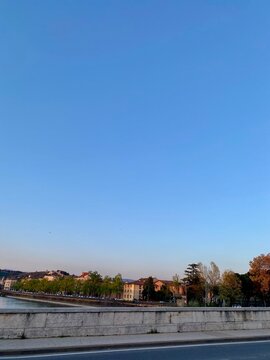 Riverside town in Italy with historic buildings and tree-lined promenade under clear blue sky, scenic European city landscape with road and bridge foreground