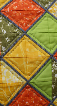 Close up on a colorful textile patchwork with diamond pattern, connected with gray zig zag stitching, representing crafting, pattern, or design