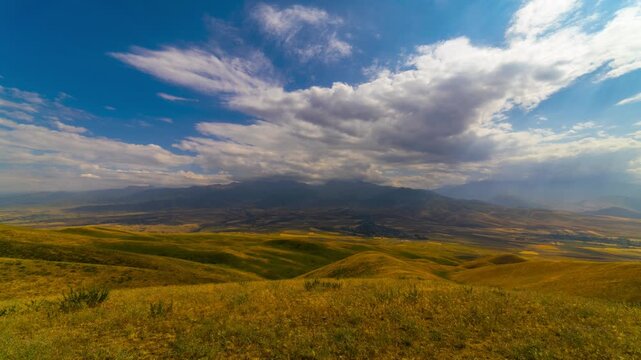 Timelapse in Kyrgyzstan: clouds slowly drift over golden rolling hills, valley village, and distant mountains with sun rays piercing dramatic clouds in the empty landscape.