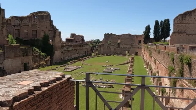 Panoramic view of ancient Roman ruins on Palatine Hill, Rome. Green lawn surrounded by crumbling brick walls, marble fragments, and towering cypress trees under blue sky.