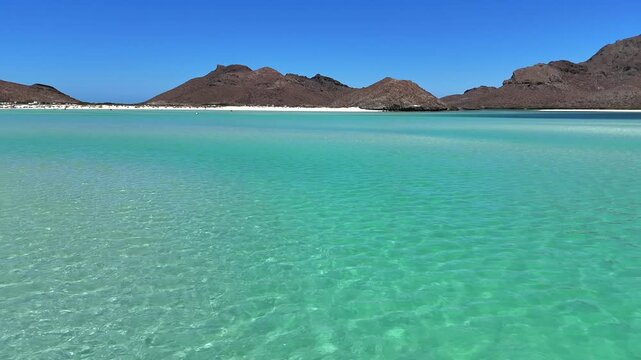 4K drone flying low over clear shallow water at Playa Balandra in La Paz, revealing the beach and mountains under bright sunlight.