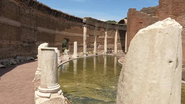 Ancient Roman ruins at Villa Adriana near Rome, Italy. Historic marble columns surround a serene reflecting pool within weathered brick walls under blue sky.