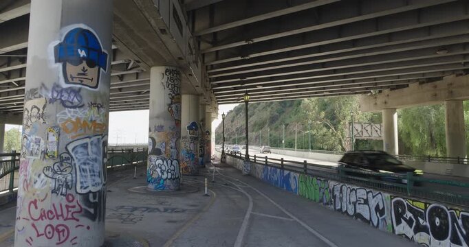 Cinematic daytime 4K 10-bit footage captured beneath the 5 and 110 freeway crossing in Los Angeles featuring large bridge support columns covered in distinct Los Angeles graffiti and street art.