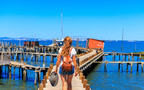 young woman with leather backpack walking on the palafitico pier of carrasqueira, authentic stilt port in comporta portugal, female traveler exploring traditional fishing village at sado estuary