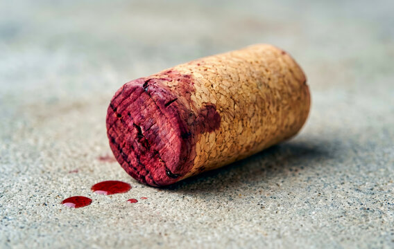 A used wine cork stained with red wine lies on a textured concrete surface with liquid drops