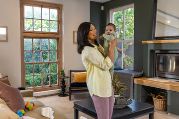 Naklejka premium Smiling mother lifting infant near face, baby reaching mouth, showing warm bond in sunlit room