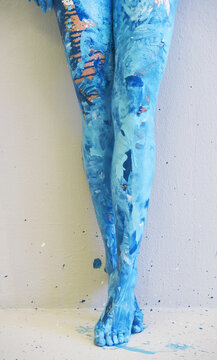 Legs covered in blue paint stand crossed against neutral wall with layered brush textures and drips creating abstract artistic body composition with strong color focus
