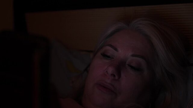Mature blonde woman lying in bed and staring at smartphone screen in complete darkness. Woman's face illuminated by cold light from mobile device during night scrolling