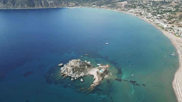 Aerial drone footage of Agios Stefanos Beach and Kastri Island in Kos, Greece. Clear turquoise water, sandy shoreline, and the small chapel on the island captured from above.