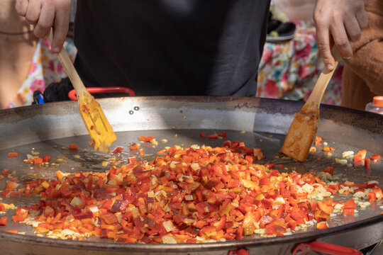 Close-up of hands cooking a sofrito of fresh vegetables in a large paella pan during an outdoor food event.
