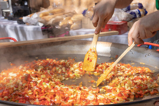 Close-up of hands cooking a sofrito of fresh vegetables in a large paella pan during an outdoor food event.