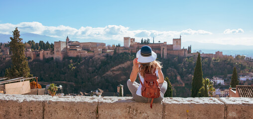 Fototapeta premium young woman with leather backpack overlooking alhambra palace in granada spain, panoramic view of the nasrid palaces and alcazaba from san nicolas viewpoint, authentic andalusian travel and heritage c
