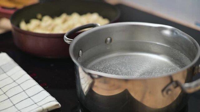 A pot filled with water is being heated while pasta is prepared in a separate pan. The kitchen setup shows essential cooking items arranged neatly on the countertop