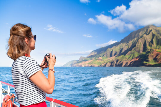 A female tourist captures the breathtaking views of the Na Pali Coast on Kauai Island from a boat as it cruises through the vibrant blue waters.