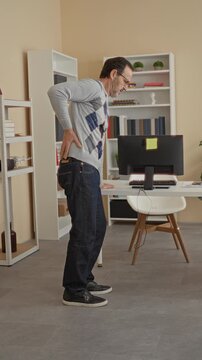 Man holding lower back and leaning on desk by computer in an office building, hand bracing back as he bends forward; workplace discomfort.