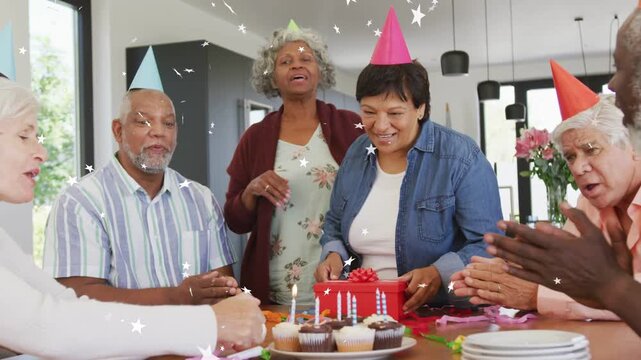 Birthday honoree shifting gift at table as candles lighting, guests singing, marketers noting stars