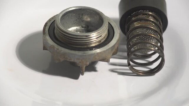Detailed macro shot of disassembled mechanical parts, featuring a metal cap, spring, and mesh filter on a white background. Perfect for automotive maintenance, industrial engineering, engine repair tu