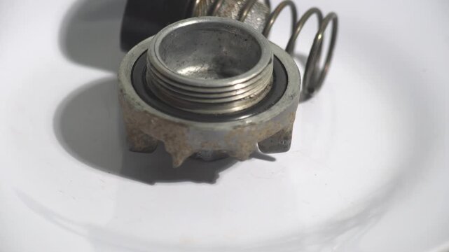 Close-up of disassembled mechanical valve components, including a threaded metal cap with rubber O-ring seal, steel spring, and mesh filter element rotating on a clean white background. Perfect for in
