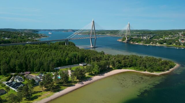 Aerial drone approach toward Uddevallabron cable-stayed bridge spanning Byfjorden fjord with forested peninsula, sandy beach and Uddevalla cityscape in view