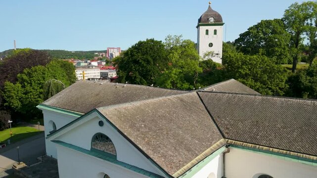 Aerial parallax drone shot orbiting white neoclassical Uddevalla Church rooftop with bell tower, clock, and city skyline visible in background