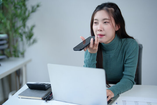 Asian businesswoman multitasking at laptop and speaking on smartphone using voice assistant or sending an audio message in a modern office creating an effective workspace