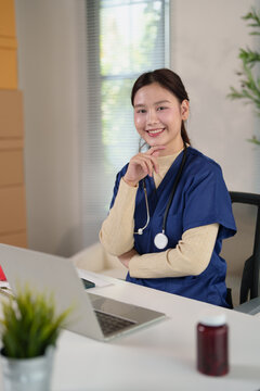 Young smiling Asian woman doctor or nurse in medical scrubs and stethoscope sitting at desk with laptop, showing professionalism, healthcare, and wellness concepts