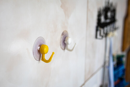 Two suction cup hooks stick to a tiled wall with shallow focus. A yellow plastic hook is sharp while a white hook blurs behind. Simple storage solution for kitchens bathrooms and rental spaces.