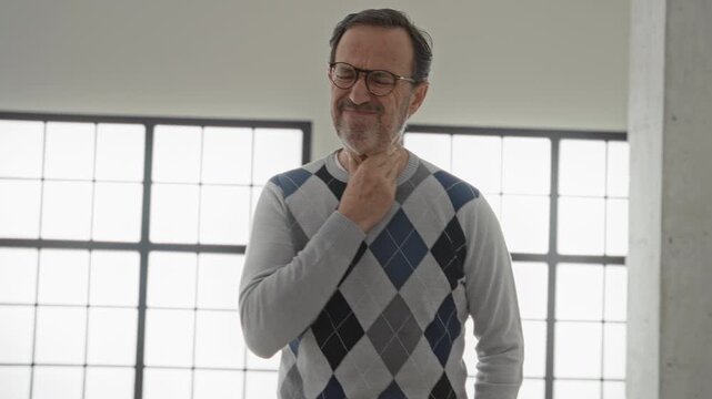 Man wearing argyle sweater and glasses clutching throat and covering mouth while coughing in studio loft with large window; sickness concern.