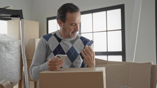 Man holding smartphone and creditcard tapping phone over cardboard box in building while unpacking and checking delivery; moving day satisfaction.