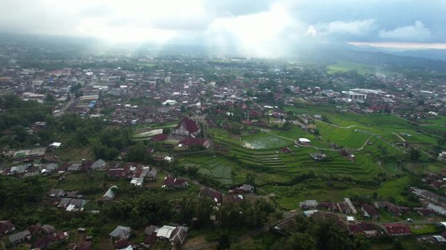 Aerial wide shot of Ruteng city in East Nusa Tenggara featuring a red-roofed church, residential houses, and green terraced rice fields under cloudy sky.
