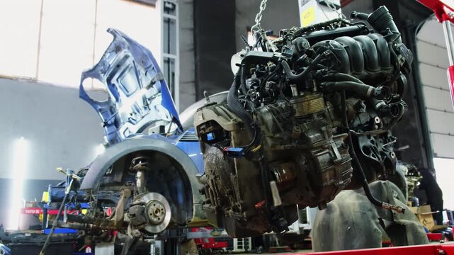 Removed engine hangs on winch chain in car service center. Technician walks past vehicle and parts performing special repair tasks