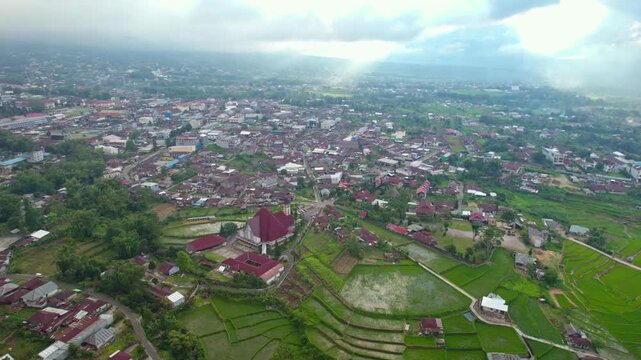 Aerial wide shot of Ruteng city in East Nusa Tenggara featuring a red-roofed church, residential houses, and green terraced rice fields under cloudy sky.
