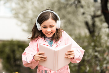 Fototapeta premium Happy 10-year-old girl using headphones to listen to an audiobook while reading in a blooming spring garden; concept of modern education and e-learning.