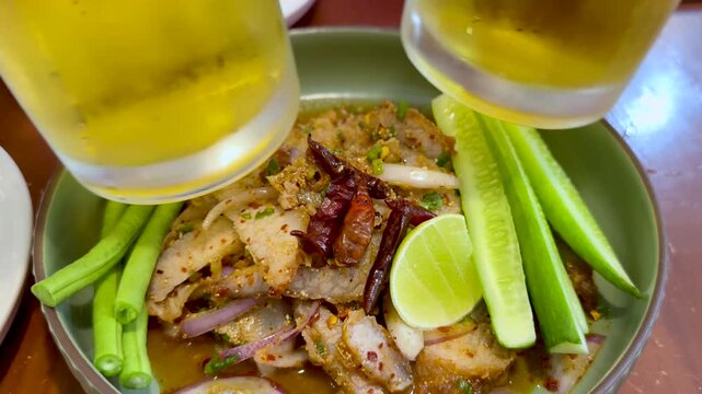 A motion footage of two hands clinking beer glasses above a vibrant Thai nam tok moo or spicy pork salad dish served with fresh vegetables.