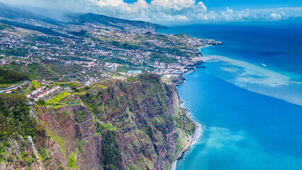 Aerial view of the dramatic cliffs of Cabo Girão, Madeira, with Funchal's urban sprawl stretching along the vibrant coastline under a clear sky. A stunning blend of nature and civilization. © Viktor