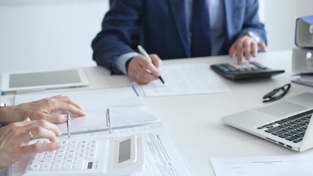 Two accountants are collaborating on a project, using a calculator and taking notes, surrounded by documents and technology in a busy office environment. Audit and taxes in business