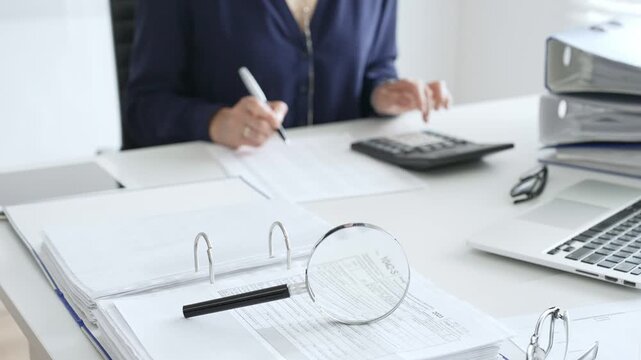 Close up of magnifying glass and calculator over financial documents opposite female accountant analyzing data, reviewing reports, binders and laptop on a white desk. Audit and taxes concept