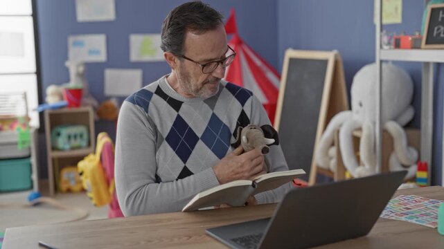 Man teacher holding stuffed elephant toy and open book, reading aloud to laptop among kindergarten toys and chalkboard in a school building; warmth connection.