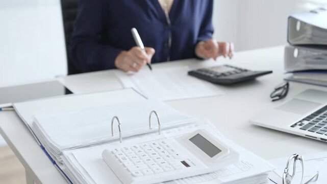 Close up of white calculator over financial documents opposite businesswoman calculating taxes at workplace, binder and glasses on desk. Audit and taxes concept
