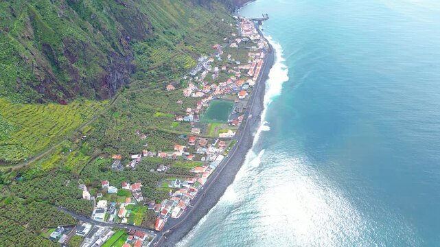 Aerial view of a vibrant Paul do Mar village on Madeira, where lush green terraced fields meet the deep blue Atlantic and a charming row of houses.