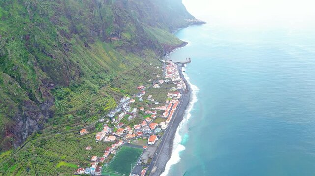 Aerial view of a vibrant Paul do Mar village on Madeira, where lush green terraced fields meet the deep blue Atlantic and a charming row of houses.