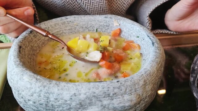 Female hand mixing chunky fish stew or soup with vegetables in rustic ceramic bowl. Traditional seafood dish served for lunch or dinner at restaurant.