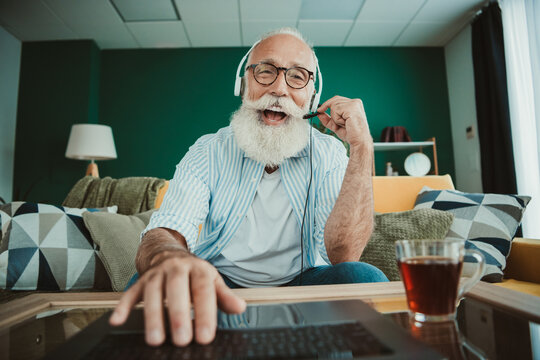 Senior man with headset sits at a laptop in a cozy living room showing happy home office and relaxation during a casual remote work day