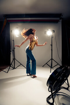 Fashion model posing dynamically in studio photoshoot with strong backlighting, wind fan movement, gold sequin top, high-waisted jeans and heels