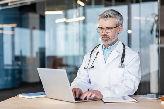 Experienced medical professional in a white lab coat and stethoscope actively using a laptop computer at a desk, focused on providing telehealth services in a modern clinic office