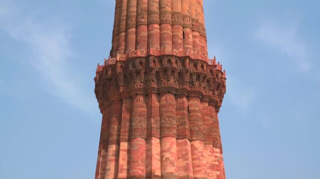 A slow tilt-up shot of the Qutb Minar minaret, showcasing its red sandstone and marble layers, intricate carvings, and balcony details against a clear sky in New Delhi.