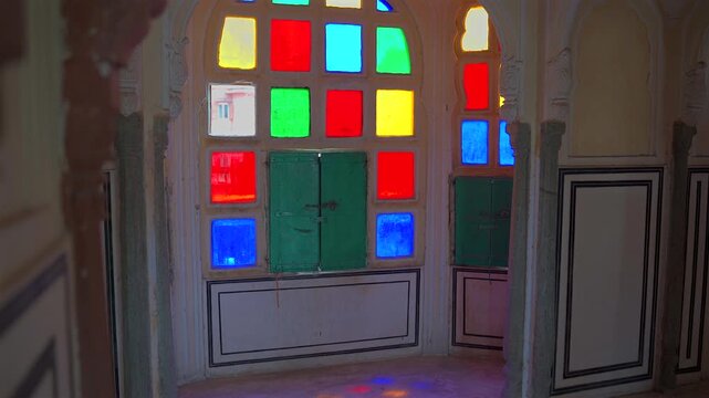 A static shot of a traditional arched window featuring vibrant red, yellow, blue, and green stained glass panes inside the Hawa Mahal, creating colorful light patterns on the walls in Jaipur.